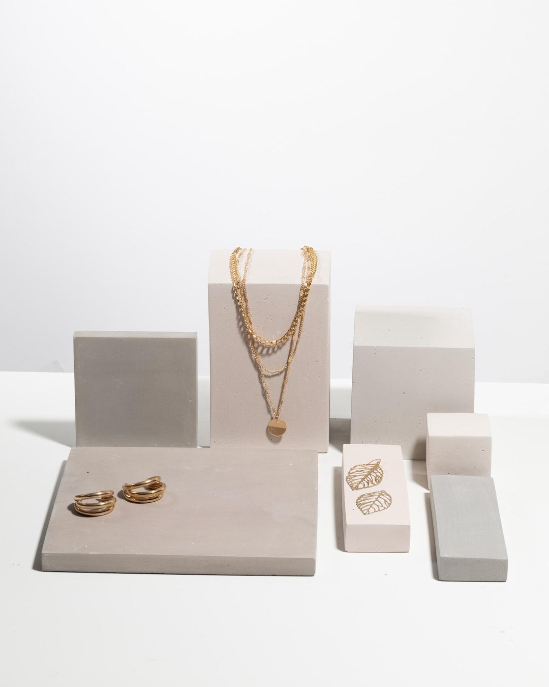 Gold layered necklace, rings and earrings displayed on taupe and grey concrete blocks
